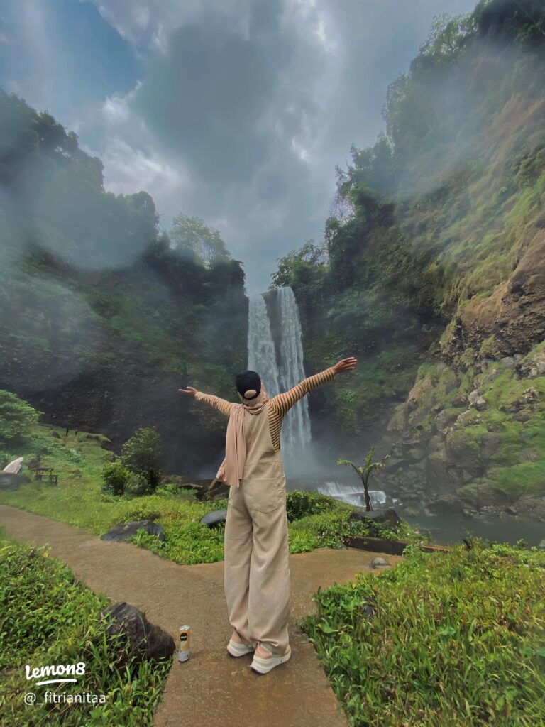 Seorang traveler berdiri menghadap air terjun besar di tengah alam hijau Indonesia